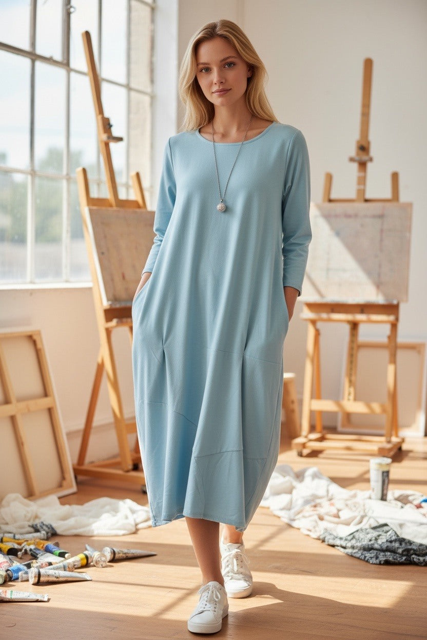 Woman in a light blue dress standing in an artist's studio with easels and art supplies.