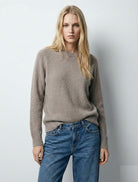 Woman wearing a taupe sweater and blue jeans against a plain background