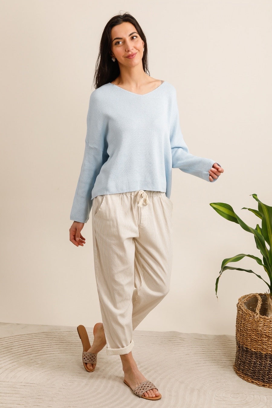 Woman wearing a light blue sweater and beige pants standing in a room with a plant and basket.