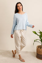 Woman wearing a light blue sweater and beige pants standing in a room with a plant and basket.