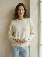 Woman wearing a cream sweater with button details against a neutral wall.