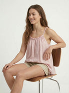 Woman sitting on a chair wearing a pink tank top and beige shorts against a white background