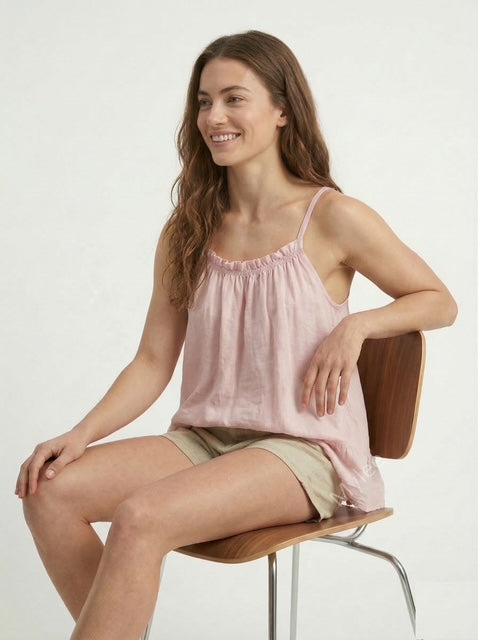 Woman sitting on a chair wearing a pink tank top and beige shorts against a white background