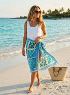 Woman on a beach wearing a white tank top and a colorful sarong, with a straw bag beside her.