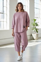 Woman wearing a pink outfit standing in a bright room with large windows.