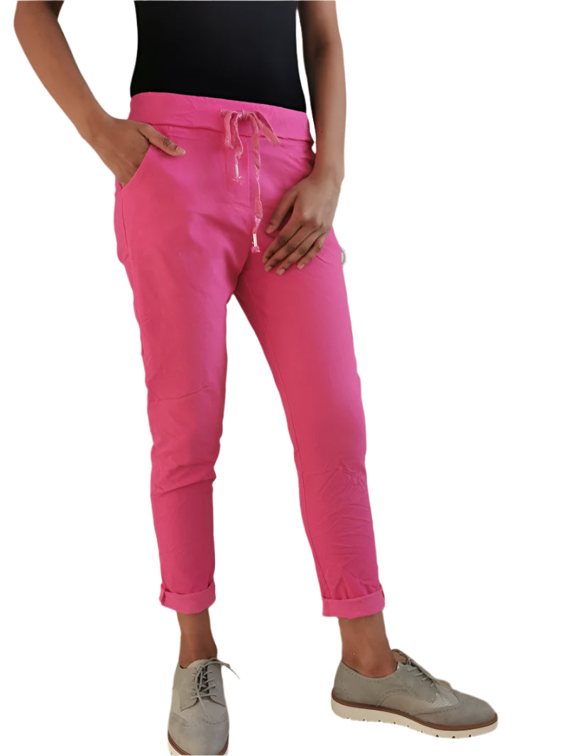 A person standing in an indoor setting wearing fuchsia Magic stretch trousers with an elasticated waist, side pockets, and white sneakers.