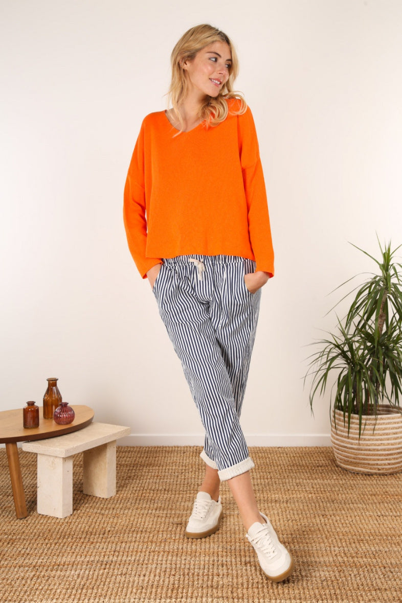 Woman wearing an orange sweater and striped pants standing in a room with a plant and small table.