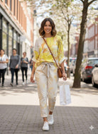 Woman walking on a city street wearing a yellow top and white pants with a brown bag.